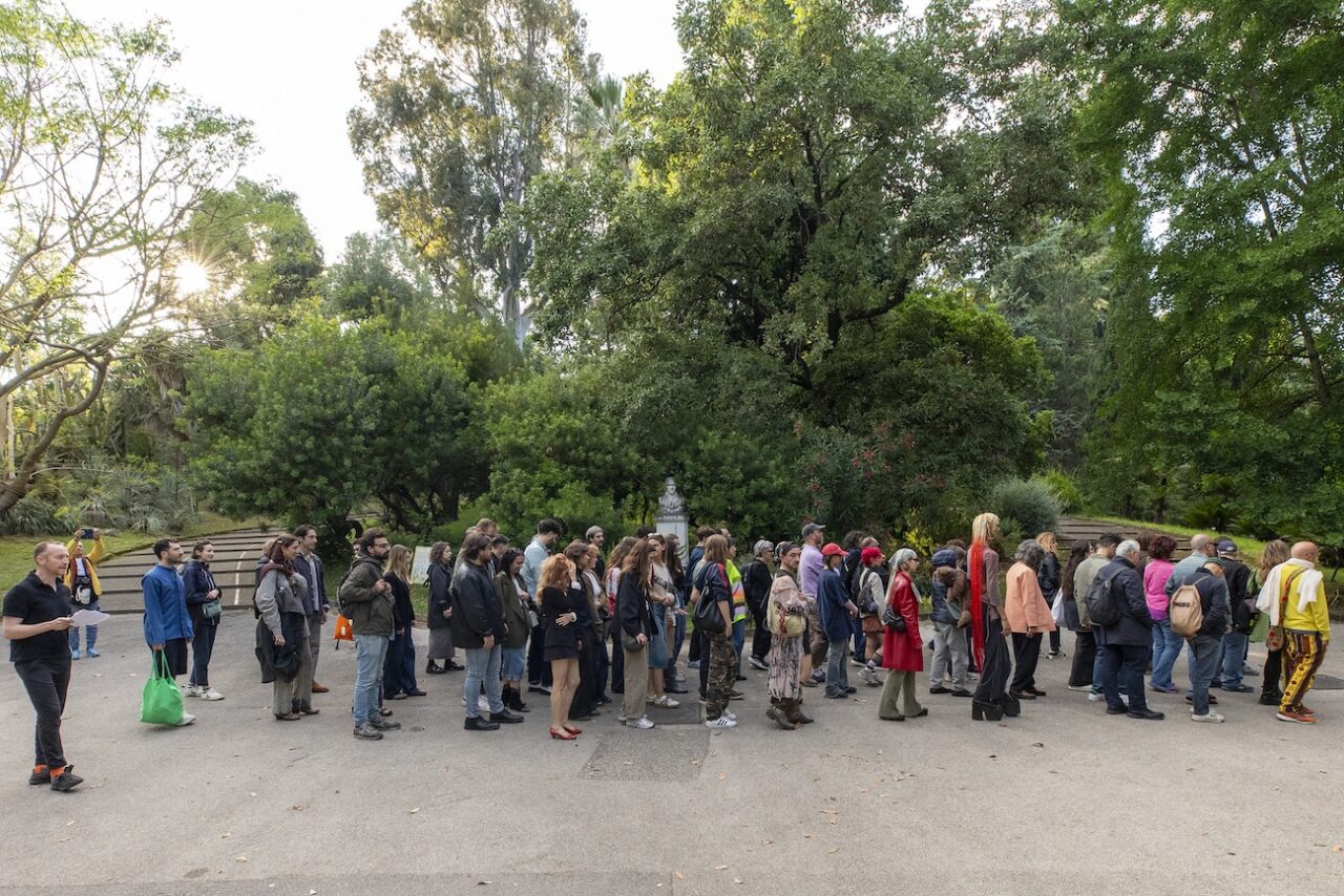 La imatge mostra un grup nombrós de persones formant una fila en un entorn enjardinat i frondós, corresponent al Jardí Botànic de Nàpols.