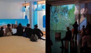 A l'esquerra, cinc persones descansen sobre una duna artificial mentre observen el documental The Terminal Beach, projectat a una pantalla LED. A la dreta, un grup de visitants recorre la sala d'exposició contemplant les projeccions del projecte Presoners de l'Amor, que es despleguen sobre diverses superfícies de l'espai.