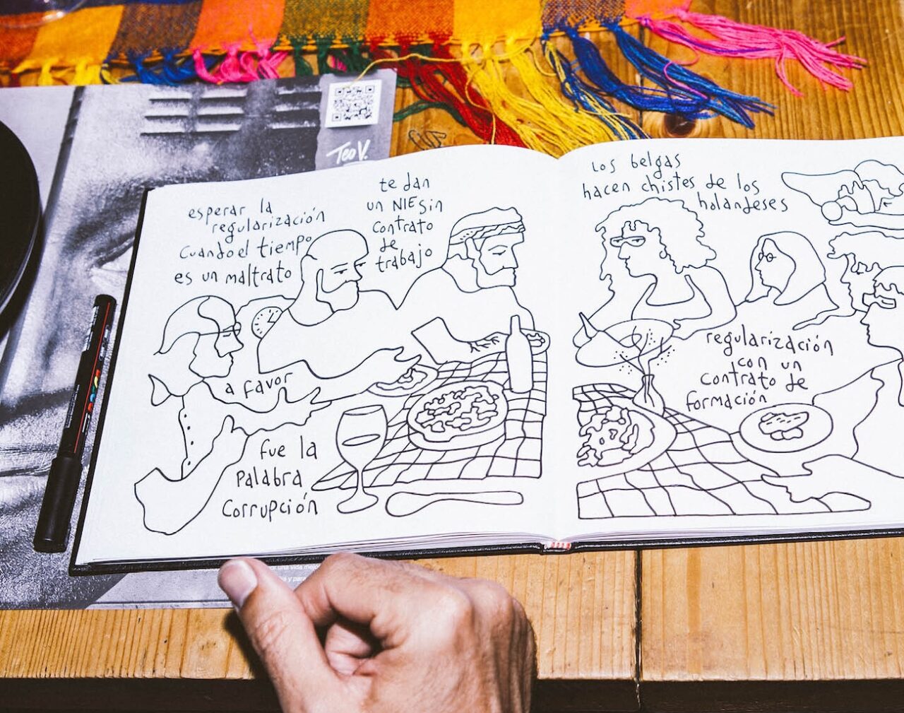 Cuaderno abierto sobre una mesa de madera con un dibujo en blanco y negro de varias personas sentadas alrededor de una mesa con comida y bebida, con frases en castellano sobre sus cabezas. Se ve una mano sosteniendo el cuaderno y un marcador a la izquierda.