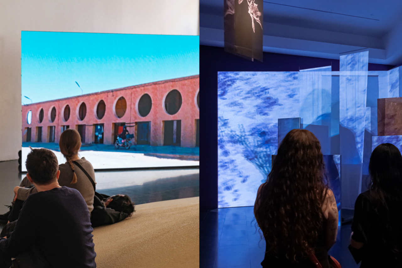 A l'esquerra, tres persones observen el documental The Terminal Beach, amb imatges d'edificacions vermelloses i geometries que irrompen al paisatge desèrtic. A la dreta, dues dones contemplen la projecció de Presoners de l'amor que, en desplegar-se sobre murs i teles suspeses, activa un joc de llums i ombres a l'espai expositiu.