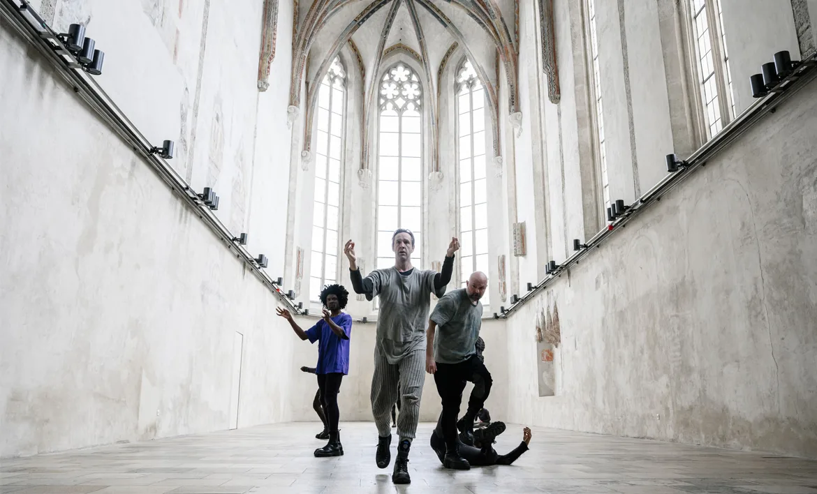 Quatre persones fan una performance dins una capella gòtica: un home al capdavant camina cap a la càmera amb els braços aixecats, una dona a l'esquerra estén els braços cap a la paret, un home a la dreta s'inclina, i una altra persona jeu a terra amb un braç aixecat.