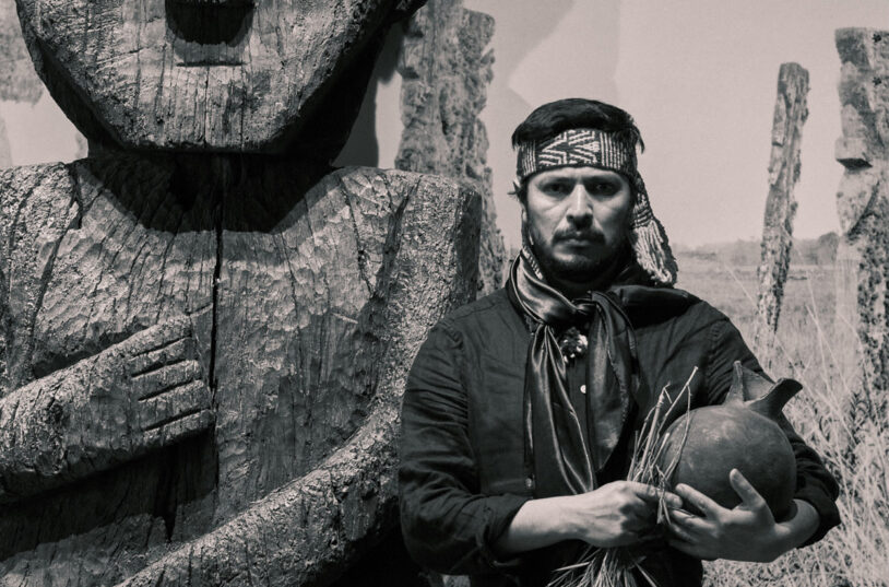 Fotografia en blanc i negre de Francisco Huichaqueo, que posa davant d'un ídol tallat en fusta, sostenint un càntir de fang a la mà esquerra i un conjunt de branques medicinals a la dreta.