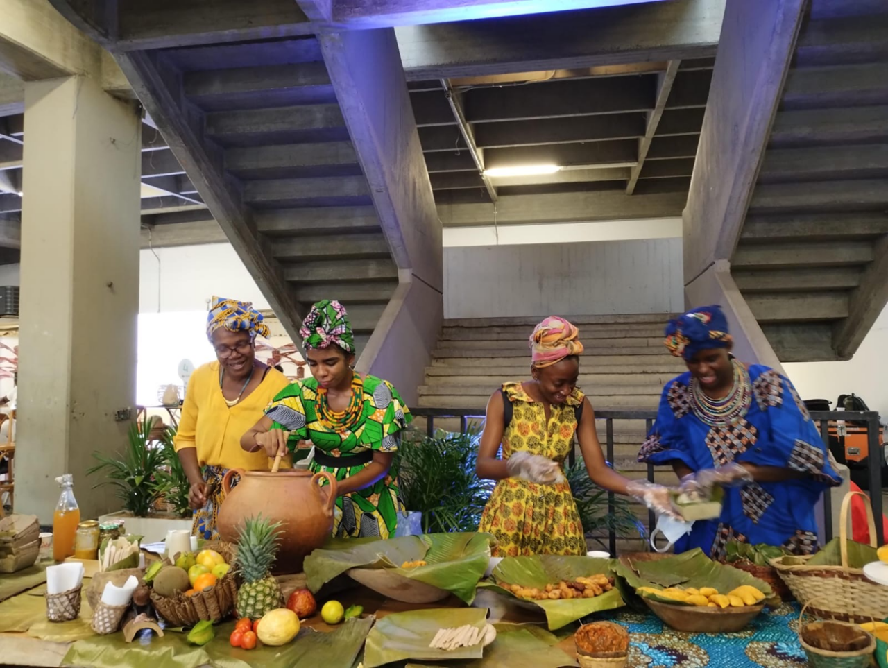 Quatre dones afrocolombianes cuinen juntes. Les dues de l’esquerra baten el contingut d’un càntir, mentre les dues de la dreta, amb guants de plàstic, preparen croquetes de plàtan. Davant seu hi ha una varietat de fruites (pinya, carambola, poma, taronja i coco) i diversos plats coberts amb fulles de plataner.