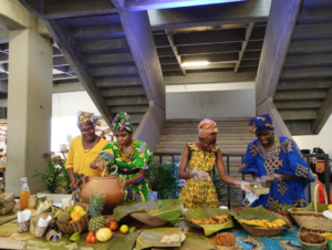 Quatre dones afrocolombianes cuinen juntes. Les dues de l’esquerra baten el contingut d’un càntir, mentre les dues de la dreta, amb guants de plàstic, preparen croquetes de plàtan. Davant seu hi ha una varietat de fruites (pinya, carambola, poma, taronja i coco) i diversos plats coberts amb fulles de plataner.