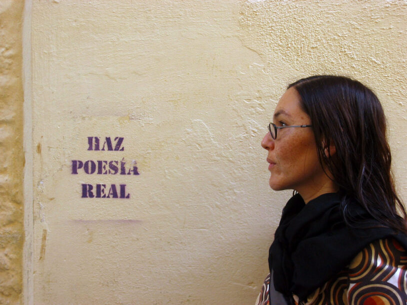 Vivian Abenshushan de perfil, mirando hacia un muro de piedra clara donde un stencil en morado reza "HAZ POESÍA REAL". Lleva gafas de montura fina, bufanda negra y una blusa estampada con círculos de colores cálidos. La composición crea un diálogo visual entre la escritora y la consigna en la pared.