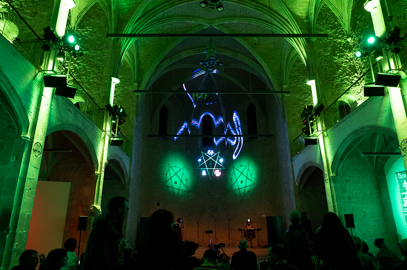 Interior de la Capella MACBA il·luminada amb llums verdes, amb públic observant un artista que actua i toca el piano.
