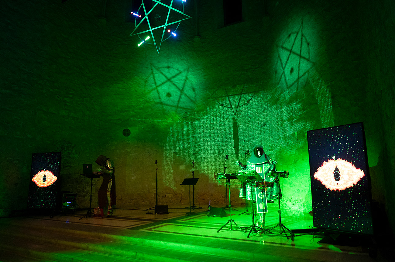 Persona disfressada manipulant un ordinador i un altra amb un teclat en un espai il·luminat de color verd amb una gran projecció a la paret.